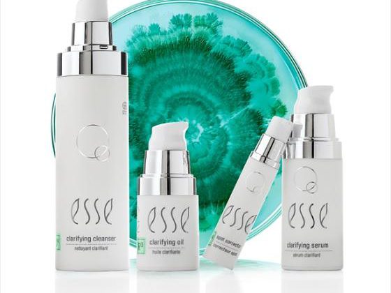 A product display featuring four white skincare bottles labeled "esse": a clarifying cleanser, clarifying oil, an eye product, and a clarifying serum. The items are arranged in front of a teal and white marbled background.