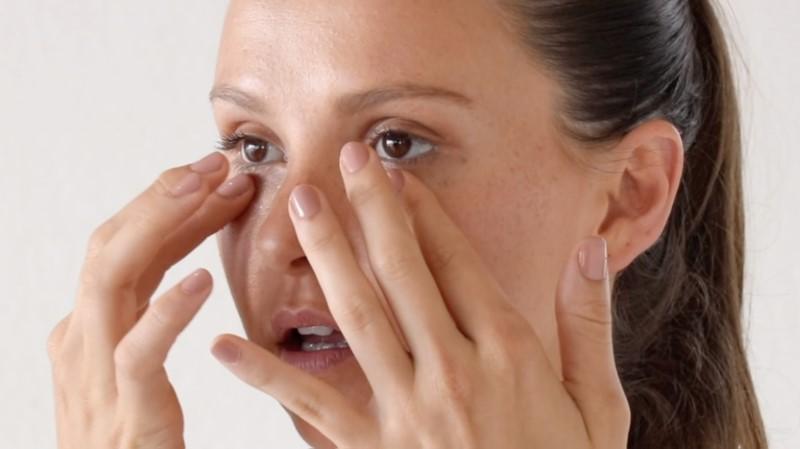 A person with dark hair in a ponytail gently touches the area under their eyes with both hands, suggesting they might be applying Esse Eye & Lip Cream or massaging the area. Their nails are neatly manicured with a light pink polish, set against a plain, light-colored background.