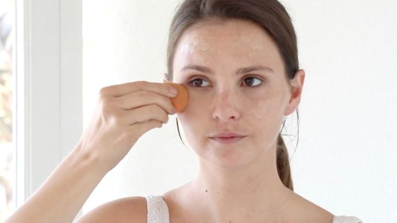 A woman with brown hair tied back uses an Esse Foundation makeup sponge to blend the foundation on her face. She is wearing a white tank top and standing against a white background.