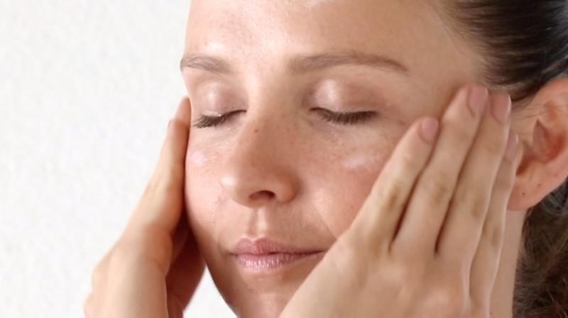 A person with closed eyes gently massages their face with both hands, focusing on the cheeks and temples using Esse Hydro Moisturiser. The skin shows a natural complexion with light freckles against a plain white background.