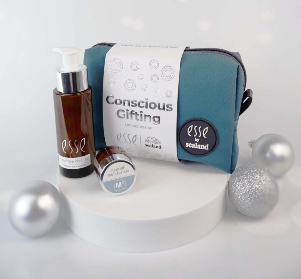The Esse Conscious Gifting set features a teal cosmetic bag with two Esse skincare products beside it, accented by silver holiday ornaments on a soft-focus backdrop.
